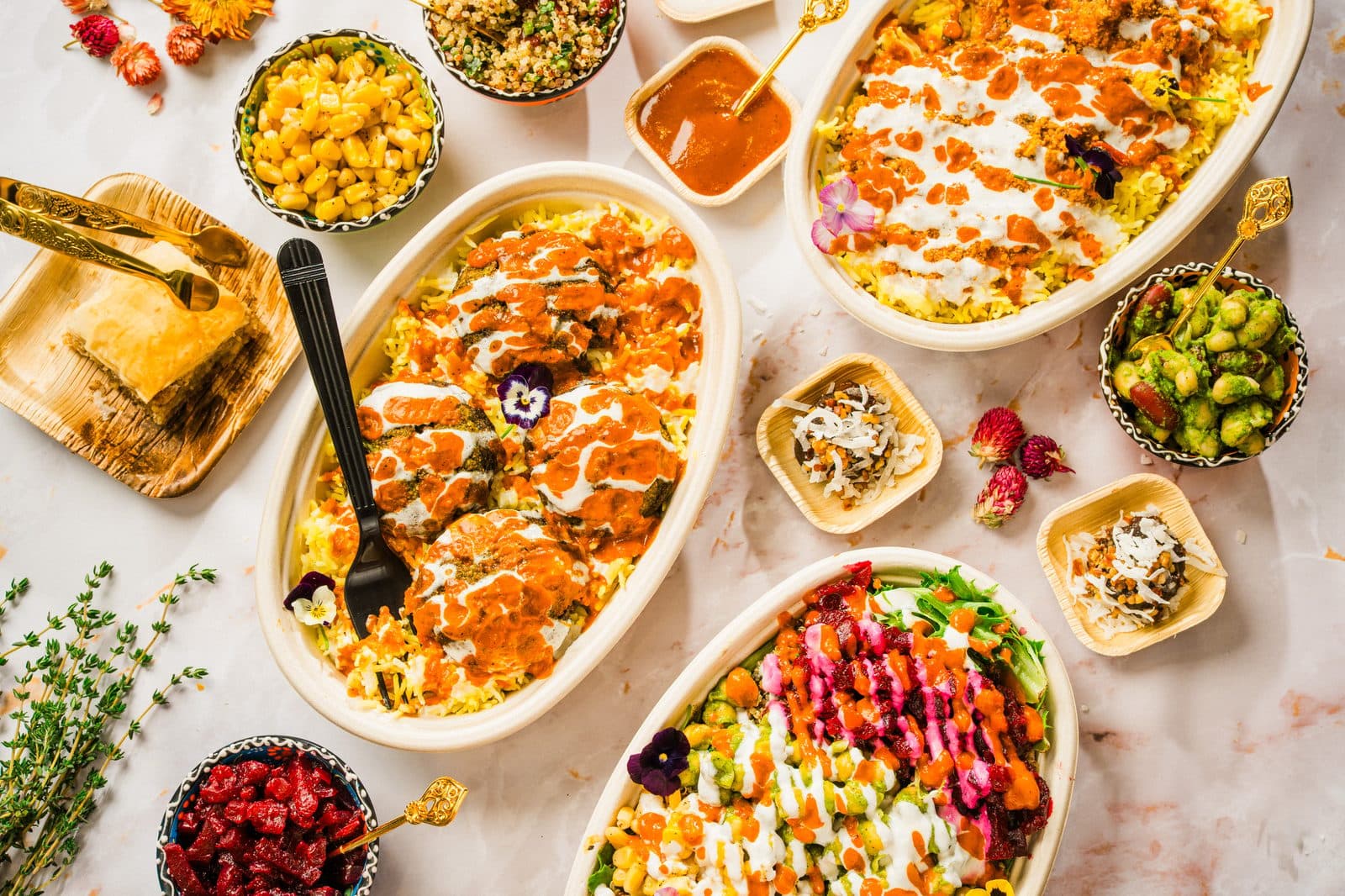 Three oval dishes of rice finished with orange and white sauce, with small bowls of sides and a square of pastry on a marble table, photographed from above