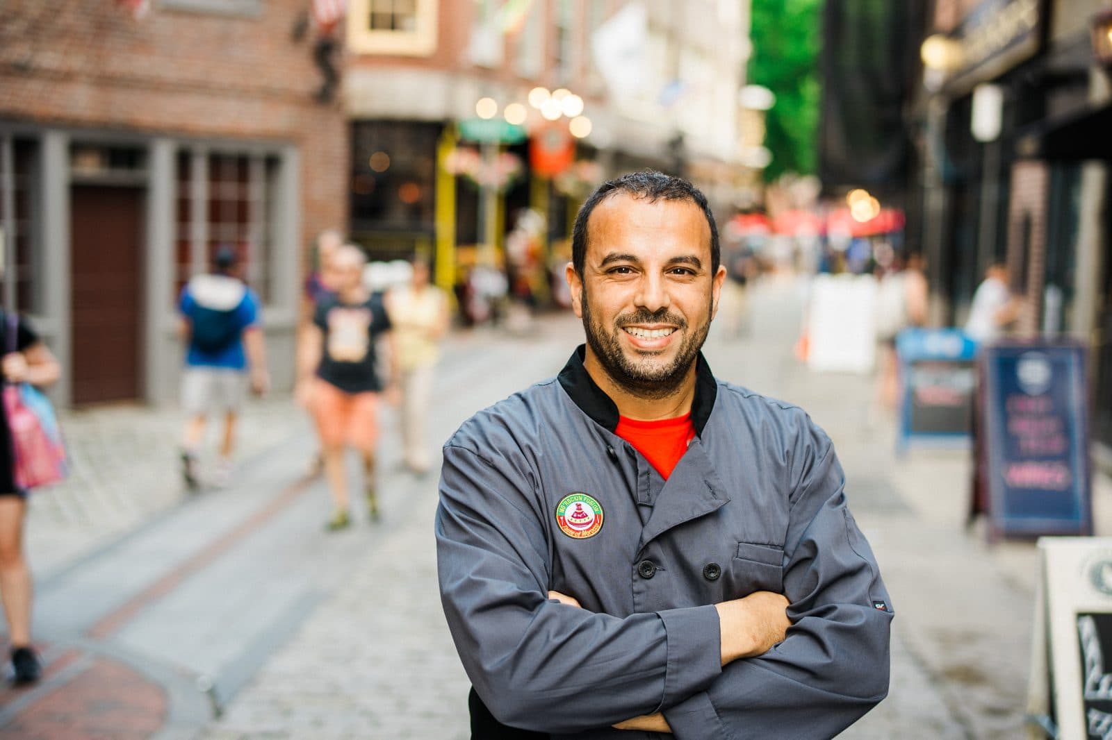 A man in a Mo'Rockin Fusion jacket standing with his arms folded on a Boston street