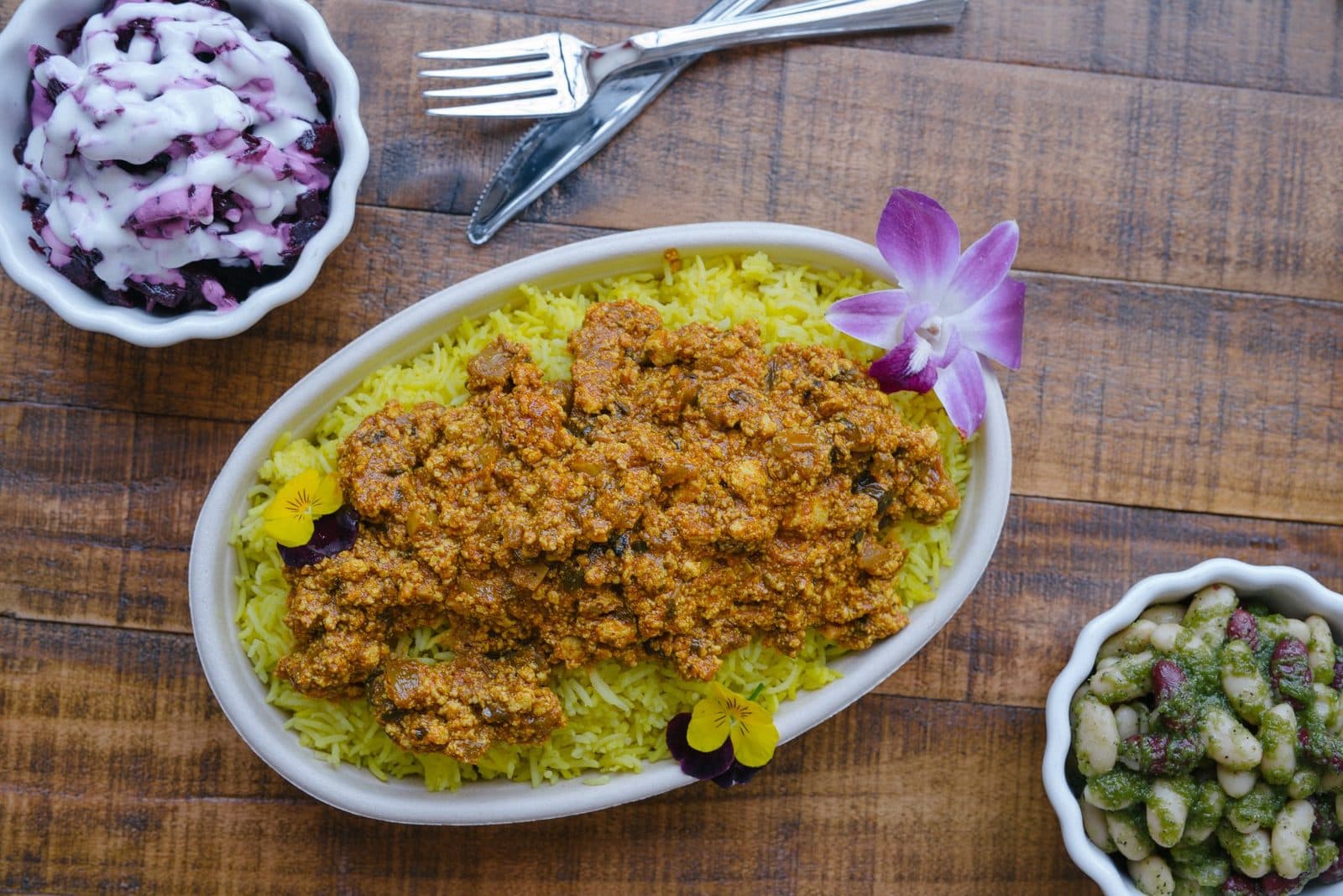 An oval dish of yellow rice topped with a spiced red mixture, with sides and a purple orchid