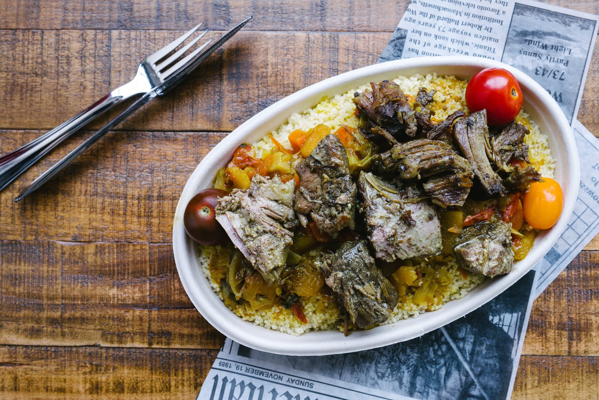 An oval dish of slow cooked meat and vegetables over couscous with cherry tomatoes, beside a knife and fork on a wooden table