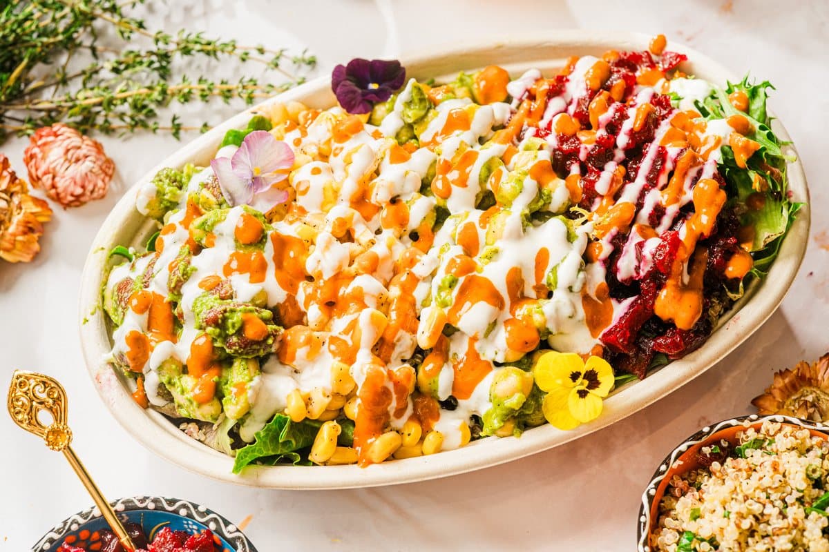 An oval bowl of greens, vegetables and beets finished with orange and white sauce, with herbs and flowers on marble