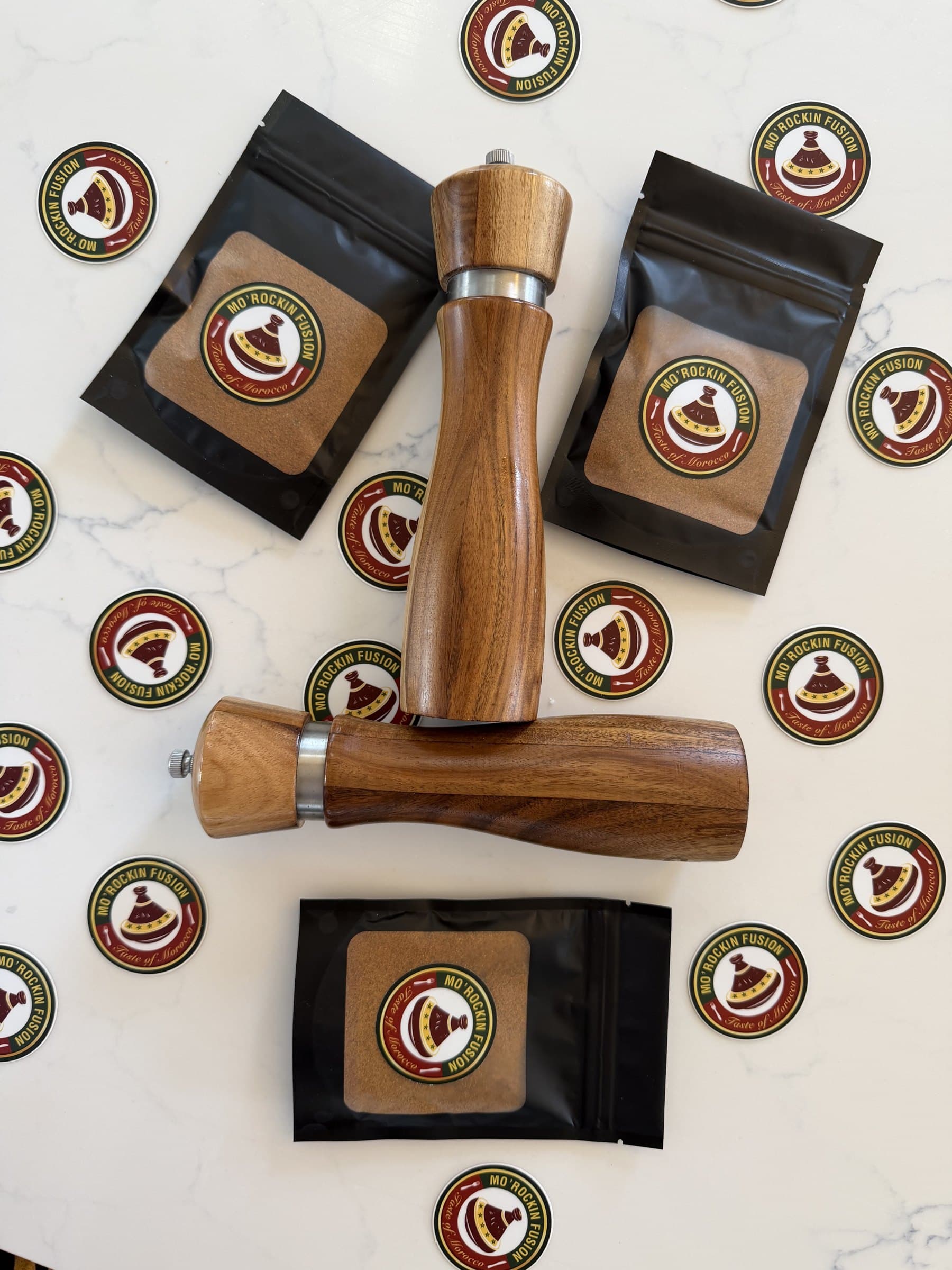 Black spice bags with the round Mo'Rockin Fusion label and two wooden grinders on white marble